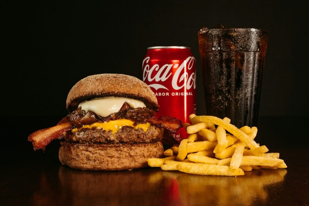 Image of a burger with bacon, fried onions, processed cheese, sauce, french fries and a can of coke. These foods contribute to subcutaneous and visceral fat accumulation.