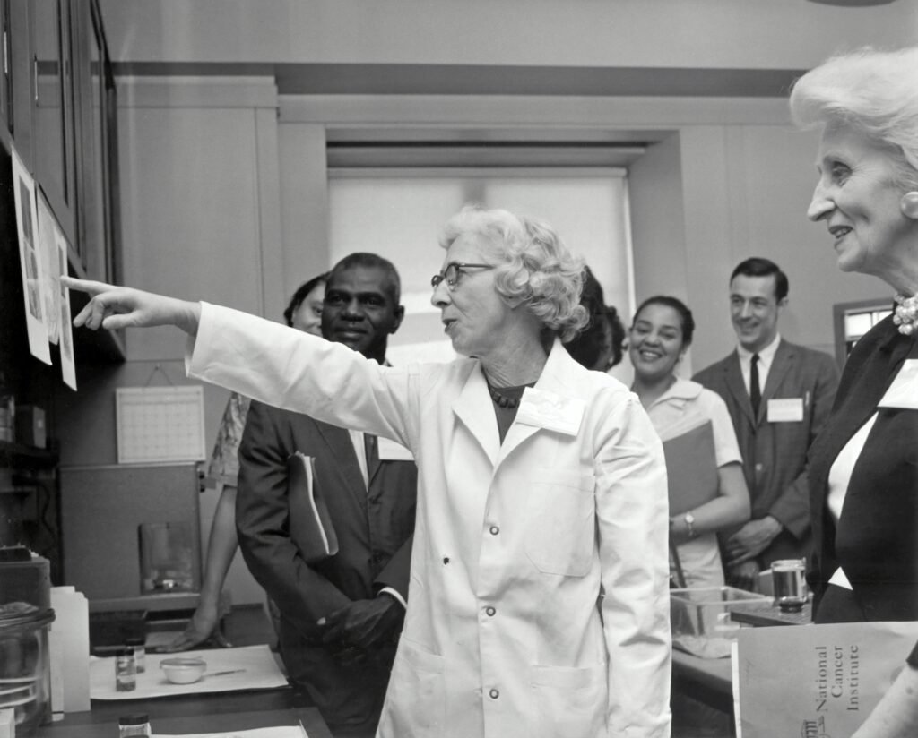 Black and white photo from what looks like the 60's with a female scientist surrounded by male scientists and male business men