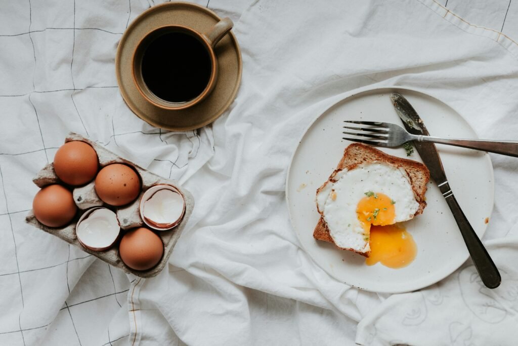 Image of 6 eggs in their carton sitting on a table cloth with a plate of toast with a cooked egg on top, and a cup of black coffee.